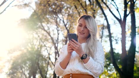 Portrait-of-girl-in-white-sweater-writing-message-on-her-smartphone-outdoors.-Woman-using-digital-gadget,-scrolls-through-social-media,-reading-news-on-app,-at-sunset.-Cute-girl-in-city-park