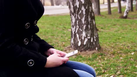 Chica-joven-usando-un-teléfono-móvil-en-un-banco-en-el-parque-de-la-ciudad