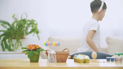 Little-nice-boy-was-sitting-at-the-table,-but-got-tired-and-decided-to-have-a-rest.-Boy-stands-out-of-the-table-with-Easter-decorations-and-is-jumping-into-a-sofa.