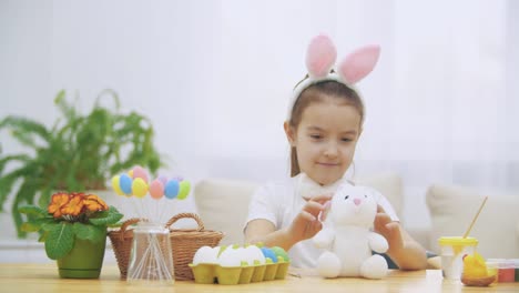 Brisk,-cute-girl-is-having-fun-painting-with-Easter-bunny,-which-has-the-same-ears,-as-she-has.-Girl-is-setting-back-white-bunny.-Girl-with-a-beauty-spot-at-her-face-and-is-smiling-widely,-sitting-at-the-wooden-table-with-Easter-decorations.