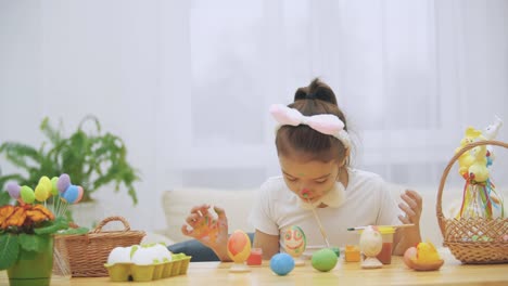 Cute-girl-is-having-fun-painting.-Adorable-girl-is-colorizing-an-Easter-egg-and-her-hands-with-a-help-of-paint-brush-in-her-mouth.-Girl-with-beauty-spots-at-her-face-and-is-looking-gently-with-cat's-whisker,-sitting-at-the-wooden-table-with-Easter-decorat