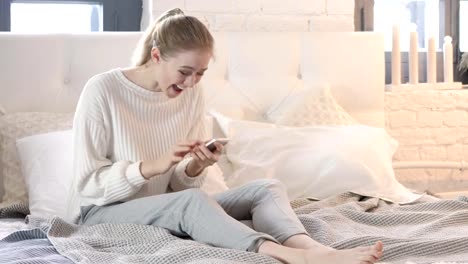 Mujer-emocionada-sentada-en-la-cama-celebrando-el-éxito-en-Smartphone