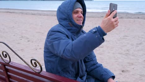 Man-blogger-in-a-blue-down-jacket-sitting-on-a-bench-on-the-sand-beach-and-talking-a-video-chat-on-mobile-phone.