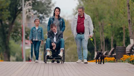 Caring-family-with-a-disabled-teenager-walks-through-the-park-on-an-autumn-afternoon.-Young-man-in-a-wheelchair-spends-the-day-in-the-park-with-a-dog-and-parents.