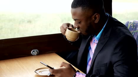 Afro-american-businessman-is-typing-a-message-on-smartphone-sitting-in-cafe.