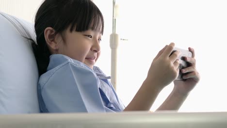 Asian-little-girl-was-lying-in-hospital-bed-and-emotion-happy-playing-video-game-online-on-smartphone.-The-sick-girl-is-a-patient-in-hospital-and-modern-medical-care.