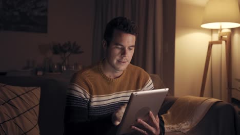 Portrait-of-a-smiling-man-sitting-on-couch-at-late-night-working-with-digital-tablet-at-home-cinematic