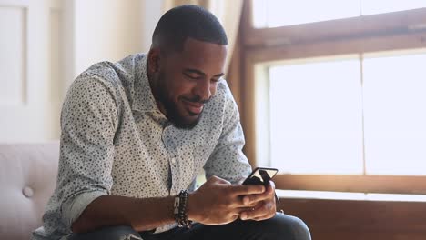 Sonriendo-hombre-afroamericano-sosteniendo-mensajes-de-texto-de-teléfono-inteligente-en-casa