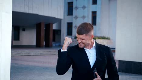 Businessman-celebrating-success.-Man-received-good-news.-Successful-Businessman-showing-Yes-gesture,-extremely-happy-about-good-news-from-smartphone.-Office-building-background.