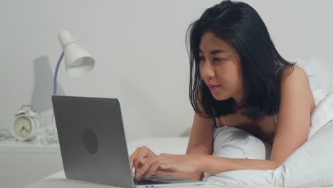 Junge-asiatische-Frau-mit-Laptop-Überprüfen-Social-Media-Gefühl-glücklich-lächeln,-während-auf-dem-Bett-liegen-duponieren,-nachdem-sie-morgens-im-Haus-aufwachen,-attraktive-thai-Mädchen-lächelnd-entspannen-im-Schlafzimmer-zu-Hause-Konzept.