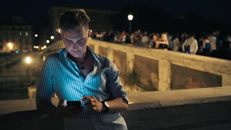 Joven-guapo-usando-su-teléfono-inteligente-por-la-noche.-Hombre-que-se-aloja-en-el-centro-de-la-ciudad-y-disfruta-de-la-vida-nocturna.-Hombre-turista-navegando-por-Internet-en-el-fondo-de-la-calle-llena-de-gente-por-la-noche.-Calle-nocturna-iluminada-llen