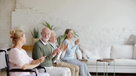 Seitenansicht-der-Gruppe-von-vier-älteren-Patienten-einschließlich-behinderter-Patienten-applaudieren-zu-Sprechen-während-der-Unterhaltungszeit-im-Pflegeheim,-konzentrieren-sich-auf-lächelnden-bärtigen-Mann-in-Brille