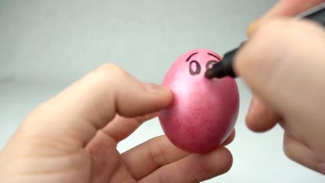 A-man-paints-with-a-felt-tip-pen-a-red-Easter-egg-draws-a-smiley-and-a-face,-a-holiday,-close-up