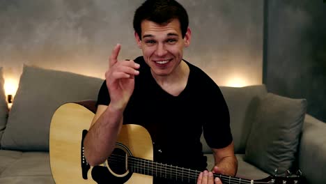 Portrait-of-short-haired-caucasian-man-in-black-T-shirt-making-video-blog-about-musical-instruments-or-recording-the-song-for-his-followers-while-sitting-on-a-couch-at-home.-Looking-to-the-camera