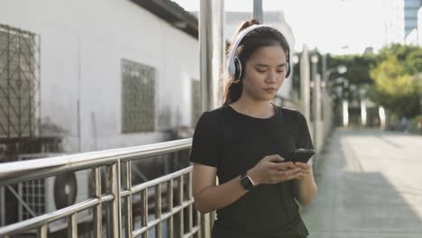 Feliz-Asia-atleta-femenina-corriendo-usando-auriculares-usando-un-teléfono-inteligente-escuchando-música-mientras-está-de-pie-fuera-en-la-ciudad-urbana.-Jogging-estilo-de-vida-saludable.