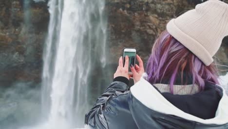 schöne-junge-Frau-in-der-Nähe-eines-Wasserfalls-in-Island,-macht-Fotos-am-Telefon,-freut-sich-und-macht-ein-Selfie