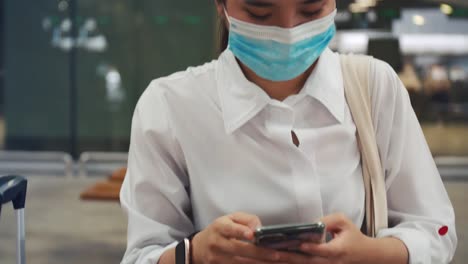 Joven-mujer-usando-protección-de-máscara-quirúrgica-y-usando-un-teléfono-inteligente-en-la-terminal-del-aeropuerto-y-mantener-una-distancia-social-durante-la-pandemia-COVID-19.