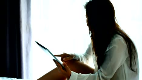 Asian-woman-use-a-tablet-on-the-bed-after-wake-up-in-weekend-mornning.Use-the-tablet-to-read-news,-watch-movies,-or-chat-in-relex-and-Communications,-social-media-concept.