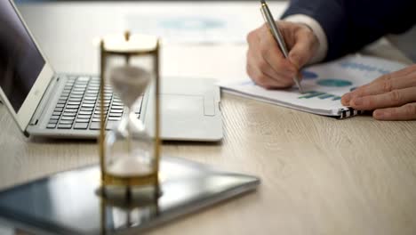 Hourglass-standing-on-table-with-sand-trickling,-man-working-with-graphs,-data