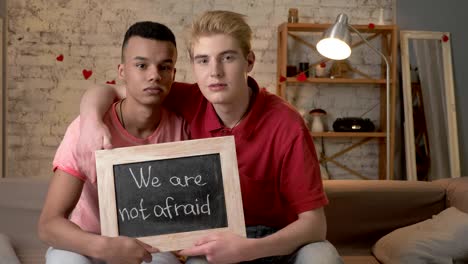 A-sad-international-gay-couple-is-sitting-on-the-couch-and-holding-a-sign.-We-are-not-afraid.-Look-at-the-camera.-Home-comfort-on-the-background.-60-fps