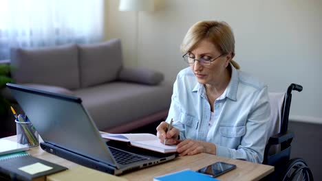 Handicapped-grandmother-making-notes-during-online-lesson-home,-senior-student