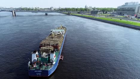 Barge-with-wood-is-pulling-up-big-blue-river-through-city-with-bridge