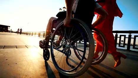 Kerl-mit-Kranken-Beinen-am-Rollstuhl-zu-Fuß-mit-Freundin-auf-der-Uferpromenade-In-den-Sommerabend