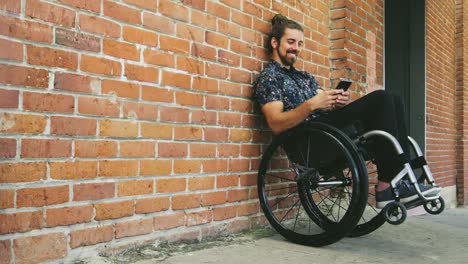 Jóvenes-discapacitados-felizes-hombre-mensajes-de-texto-de-teléfono-en-silla-de-ruedas-inclinada-contra-la-pared-de-ladrillo