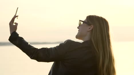 Eine-Nahaufnahme-von-einem-schönen-Mädchen-sitzen-auf-einem-Steg-am-Meer-weht-einen-Kuss-im-Sonnenuntergang-eine-Instagram-Storry-Selfie,-dabei