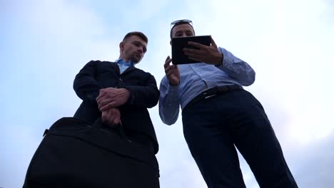 Young-businessman-showing-a-presentation-on-screen-of-tablet-pc-to-his-colleague-in-city.-Successful-entrepreneurs-working-on-digital-tablet-with-blue-sky-at-background.-Low-angle-view-Slow-motion