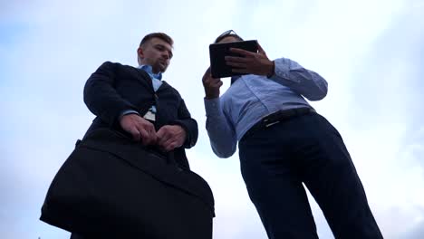 Two-young-businessmen-talking-and-using-tablet-pc-in-city.-Handsome-coworkers-discussing-and-working-on-digital-tablet-with-sky-at-background.-Colleagues-applying-mobile-technology.-Low-angle-view