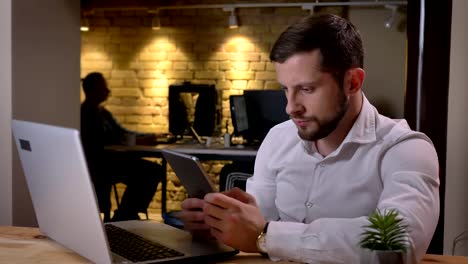 Closeup-portrait-of-young-caucasian-businessman-typing-on-the-tablet-in-front-of-the-laptop-in-the-office