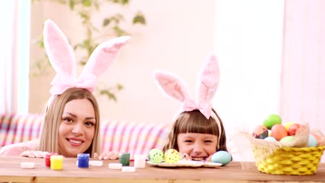 mother-and-daughter-in-headbands-with-ears-peek-out-from-behind-the-table-and-smile-widely,-on-the-table-are-paint-and-Easter-eggs.