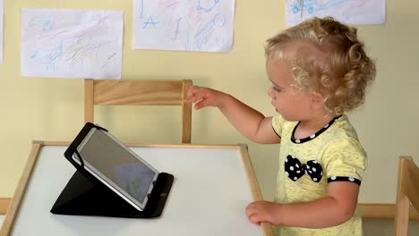 niedliche-Baby-Mädchen-mit-Tablet-Computer-sitzen-in-der-Nähe-von-einem-kleinen-Tisch.