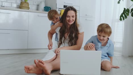 Moderne-Technik.-Moderne-Wohnung-liebende-Mutter-und-zwei-kleine-Söhne,-die-auf-dem-Boden-im-Wohnzimmer-sitzen,-schauen-auf-den-Laptop-Bildschirm.-Kinder-mit-Mama-spielen-auf-einem-Laptop