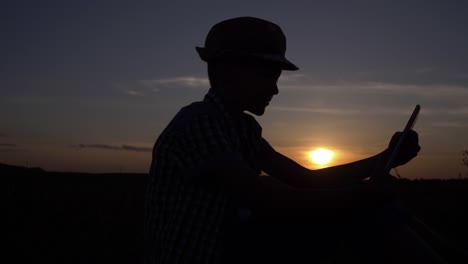 silueta-de-un-niño-utiliza-una-tableta-al-atardecer-en-el-campo,-Lee-algo-en-la-tableta,-al-aire-libre