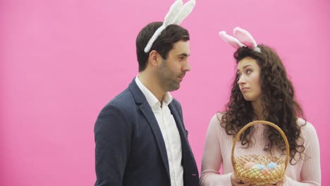 Young-beautiful-couple-standing-on-a-pink-background.-With-hackneyed-ears-on-the-head.-During-this-man-puts-a-yellow-egg,-which-was-missing-his-wife-in-a-basket.-Gently-kissing-smile.