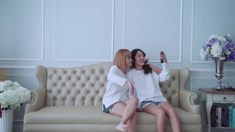 Happy-couple-friend-sitting-on-sofa-hug-and-using-phone-taking-selfie-together-at-home.-Friend-and-lesbian-concept.