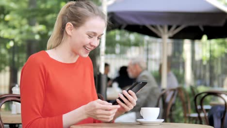 Junge-Frau-bejubelt-Erfolg-auf-Smartphone-Sitting-in-Cafe-Terrasse