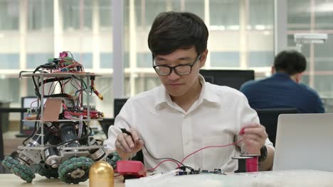 Young-asian-electronics-development-engineers-works-with-robot,-measuring-the-signal-in-the-electrical-circuits-of-robotics-prototype-in-workshop.-People-with-technology-or-innovation-concept.