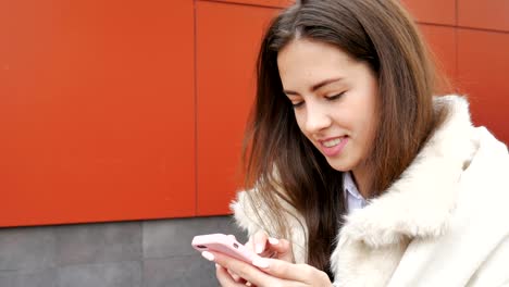 A-young-attractive-girl-stands-at-the-wall-of-a-building-and-pokes-a-finger-into-a-mobile-phone.