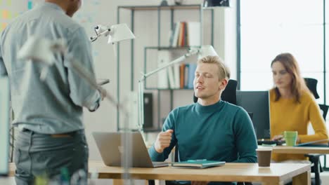 Handsome-IT-Specialist-Sitting-at-His-Desk-arbeitet-an-einem-Laptop,-spricht-mit-professionellem-Projektmanager.-Vielfältiges-Team-von-Young-Professionals,-die-im-Büro-arbeiten