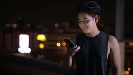 Porträt-von-jungen-glücklichen-asiatischen-Mann-mit-Telefon-im-Freien-in-der-Nacht