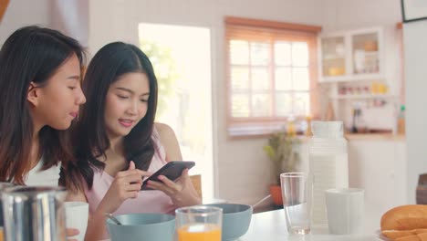 Asian-Lesbian-couple-using-mobile-phone-check-news-while-has-breakfast-in-kitchen-at-home.