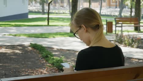 Glückliche-Mädchen-Student-mit-einem-Smartphone-in-einem-Stadtpark-sitzen-auf-einer-Bank,-junge-Frau-lächelnd-verwenden-Anwendung,-lange-blonde-Haare.