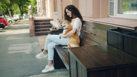 Lächelnde-Mädchen-mit-Smartphone-und-umarmung-Shiba-inu-Hund-im-Freien-im-Café