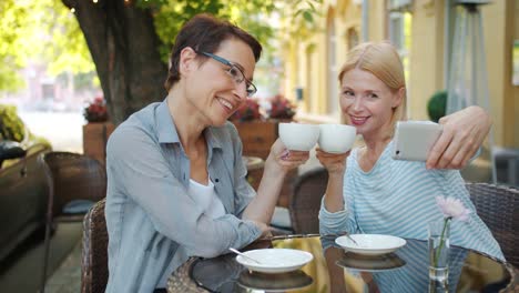 Mature-ladies-taking-selfie-with-cups-in-open-air-cafe-holding-coffee-smiling