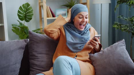 Fröhliche-Nah-Ost-Mädchen-in-Hijab-mit-Smartphone-lachen-Spaß-zu-Hause