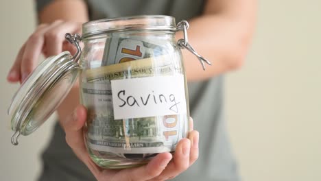 Woman-putting-paper-dollar-bill-in-a-glass-piggy-bank-with-inscription-Saving.-Save-money-concept