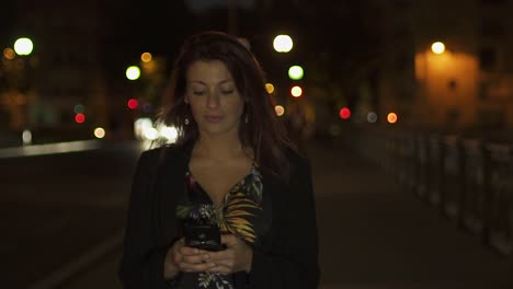 Beautiful-Attractive-caucasian-modern-woman-wearing-flower-dress,-black-jacket-and-red-hair-walking-through-the-street-and-writing-a-text-message-on-her-smartphone-by-night.-Paris-4K-UHD.-Slow-Motion.